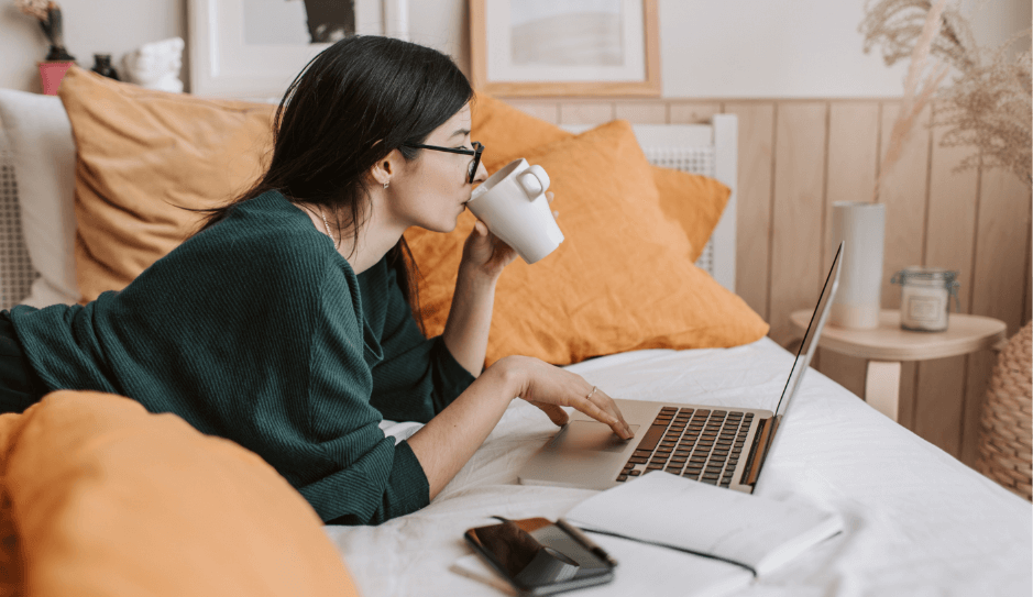 Woman working on bed on laptop drinking from her mug